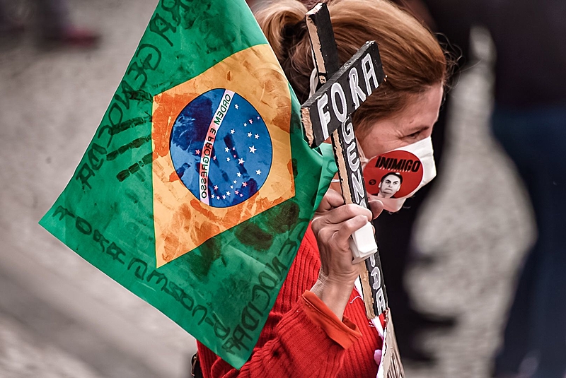 Mulher com máscara segurando bandeira do brasil e crucifixo escrito: Fora Genocidas